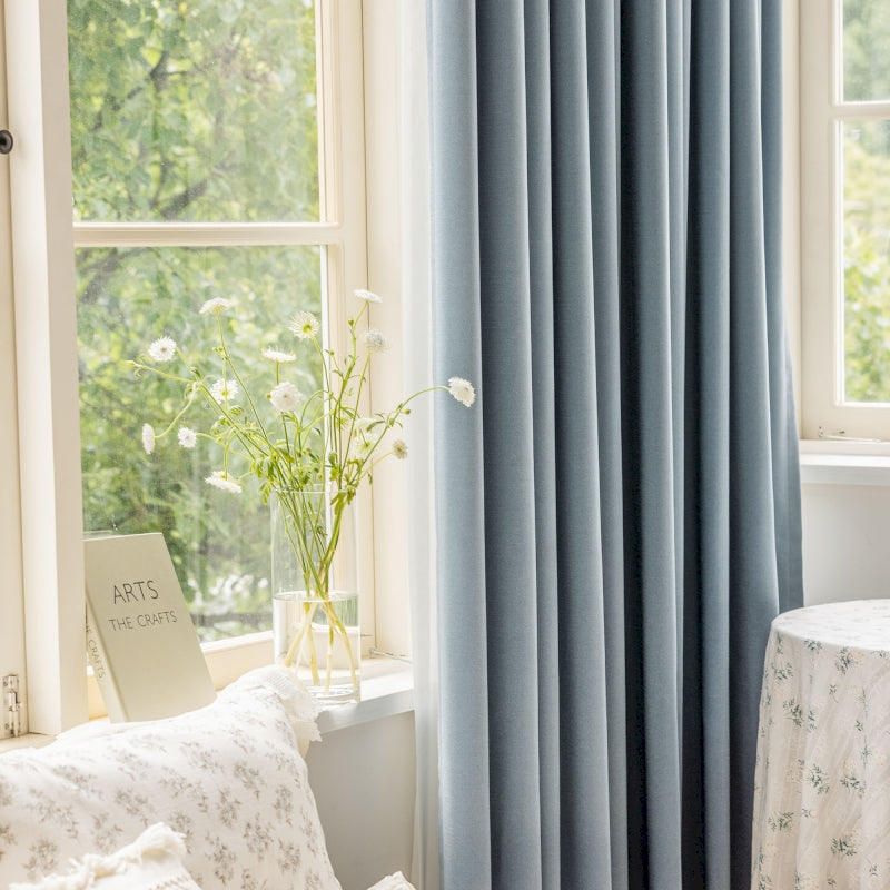 a window with a blue curtain and a white table with a vase of flowers